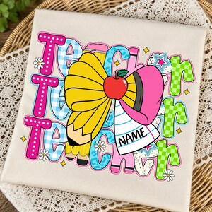 May include: A cream-coloured t-shirt featuring a colourful graphic with the word TEACHER repeated in various patterns like pink polka dots, blue gingham, and green checkers. A large yellow pencil bow with a red apple and a white name tag is centred.