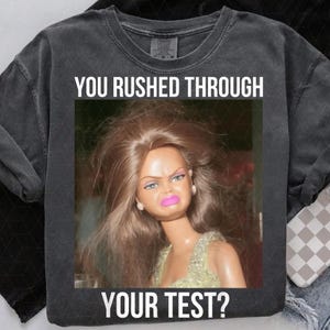 May include: Dark gray sweatshirt with the text "YOU RUSHED THROUGH YOUR TEST?" above and below a photo of a doll with a displeased expression. The doll has long brown hair, pink lipstick, and is wearing a light green dress.