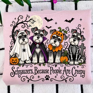Op de afbeelding: Een lichtroze sweatshirt met een afbeelding van vier Schnauzer-honden in Halloween-kostuums, waaronder een geest, vampier, pompoen en skelet. De tekst luidt "Schnauzers, Because People Are Creepy", omringd door vleermuizen, een maan en bomen.