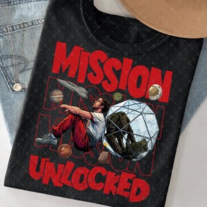 May include: Black graphic t-shirt with the words "MISSION UNLOCKED" in red. The design features a man in red pants and a white shirt, with space-themed illustrations including planets, a spaceship, and a geometric structure.