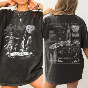 Puede incluir: Camiseta gris oscuro con un diseño frontal de Artemisa con arco y flecha, un cohete y el texto "First of Her Kind". La parte trasera de la camiseta muestra un diagrama de la misión Artemis II a la luna.