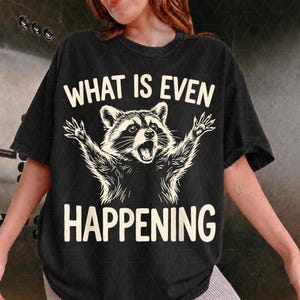 Op de afbeelding: Een zwart t-shirt met korte mouwen en een afbeelding van een wasbeer met de armen in de lucht. Boven de wasbeer staat de tekst WHAT IS EVEN en onder de wasbeer staat de tekst HAPPENING. Het ontwerp is gedrukt in een crèmekleur.