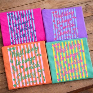 May include: Four colorful t-shirts with the text "Most likely to binge watch shows all summer." Each shirt is a different color: pink, purple, orange, and teal. The text is in a script font with vertical stripes and summer-themed graphics.
