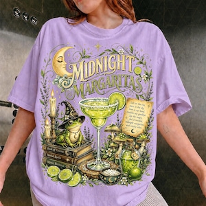 以下が含まれることがあります： 「Midnight Margaritas」の文字がプリントされたライトパープルのTシャツ。魔女の帽子をかぶって本の上に座るカエル、マルガリータグラス、キャンドル、キノコ、三日月、そして文字が書かれた羊皮紙の巻物が描かれたグラフィックデザインが特徴です。
