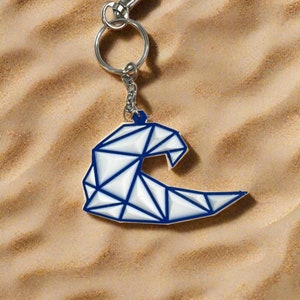 May include: A silver key chain with a geometric wave charm. The wave is white with blue lines and is on a sandy background.