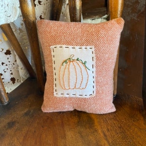 May include: A small, orange pillow with a white embroidered pumpkin design. The pillow is sitting on a wooden chair.