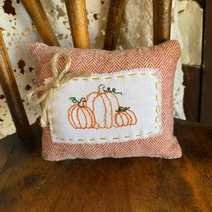 May include: A small, red and white pillow with a stitched design of three pumpkins. The pillow has a brown bow on top.