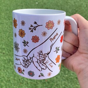 May include: White ceramic mug with a floral design and three handprints. The handprints are connected and labeled "Mama", "Jaxon", and "Evie".