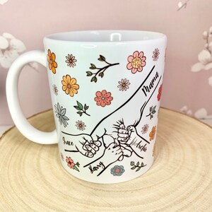 May include: White ceramic mug with a floral design and handprints with the names "Mama", "Grace", "Harry", and "Archie" written on it.