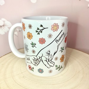 May include: White ceramic mug with a floral design and two hands holding each other. The text "Mummy" and "Jack" are written on the mug.