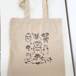 May include: Beige tote bag with black line art illustrations of wedding-themed elements. Designs include bows, roses, cake, champagne glasses, doves, and the text "Bride Era", "She's Giving Wife", "Est. 2026", and "Future Mrs."