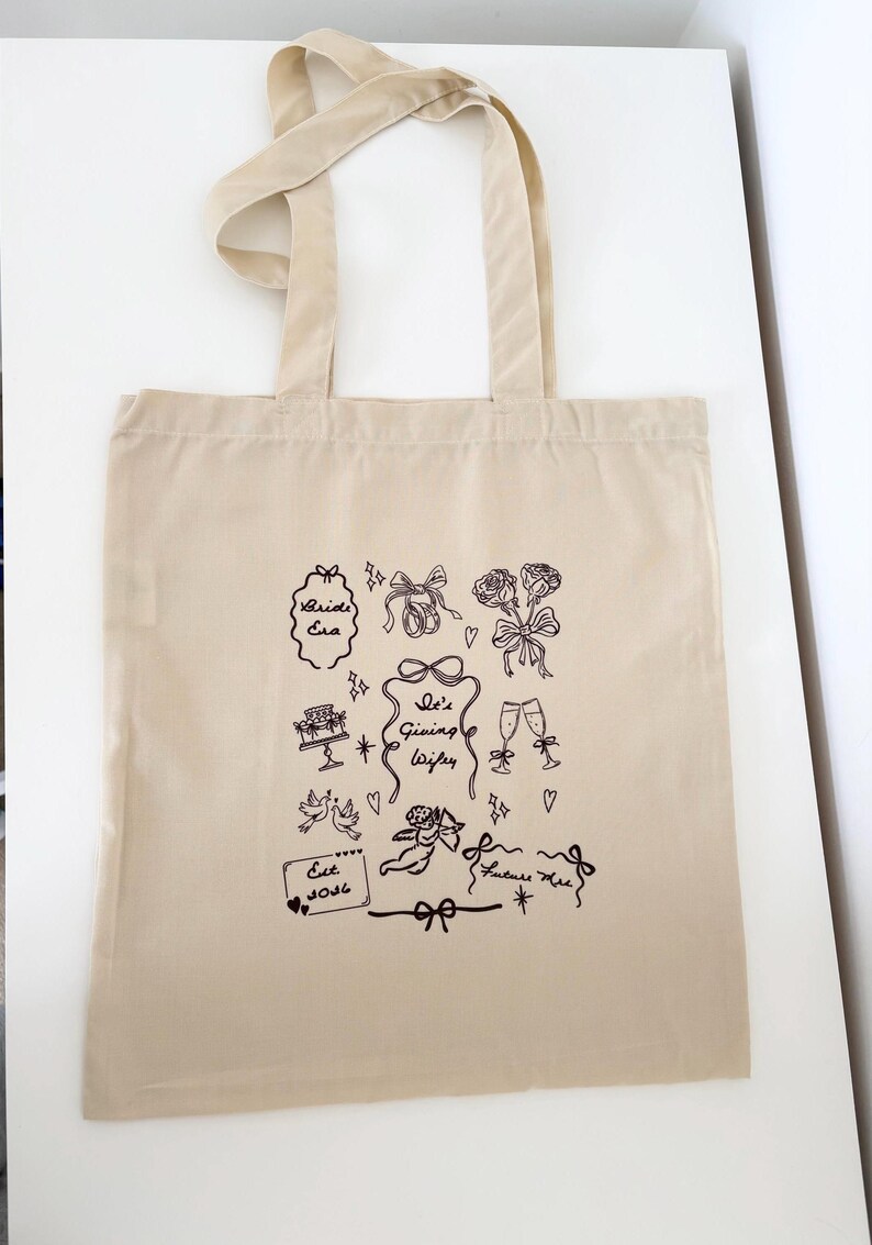 May include: Beige tote bag with black line art illustrations of wedding-themed elements. Designs include bows, roses, cake, champagne glasses, doves, and the text "Bride Era", "She's Giving Wife", "Est. 2026", and "Future Mrs."