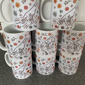 May include: Set of nine white ceramic mugs with floral designs and hand-drawn illustrations of hands holding each other. Each mug has a different name written on it, including "Mama", "Mummy", "Mum", and "Nana".