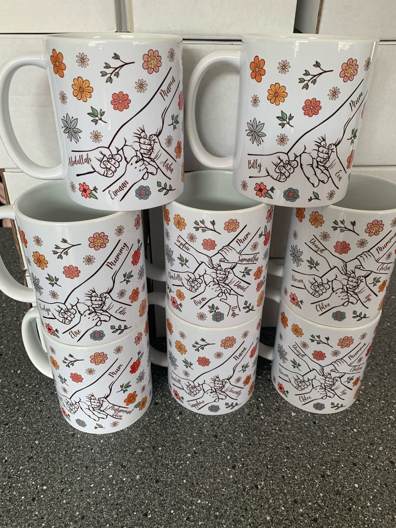 May include: Set of nine white ceramic mugs with floral designs and hand-drawn illustrations of hands holding each other. Each mug has a different name written on it, including "Mama", "Mummy", "Mum", and "Nana".