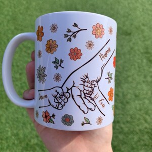 May include: White ceramic mug with a floral design and three handprints holding each other. The handprints are labeled "Mama", "Jaxon", and "Evie".
