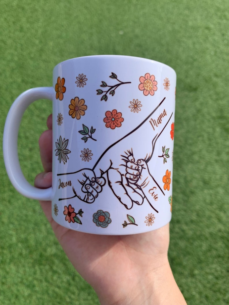 May include: White ceramic mug with a floral design and three handprints holding each other. The handprints are labeled "Mama", "Jaxon", and "Evie".