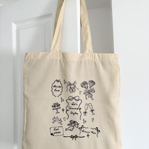 May include: Beige canvas tote bag with black line art illustrations of bows, roses, champagne flutes, and the text "Bride Era" and "It's Giving Wifey". The bag is hanging on a white door.