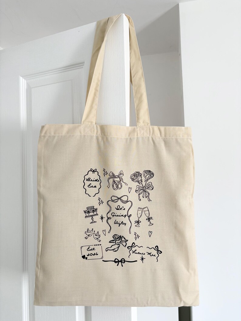 May include: Beige canvas tote bag with black line art illustrations of bows, roses, champagne flutes, and the text "Bride Era" and "It's Giving Wifey". The bag is hanging on a white door.
