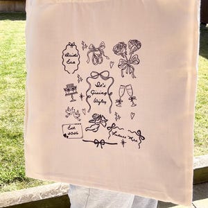 May include: Cream-colored tote bag with black line art illustrations. The design includes text that says "Bride Era", "She's Giving Wifey", and "Est. 2026". Other illustrations include bows, roses, champagne glasses, and a cake.