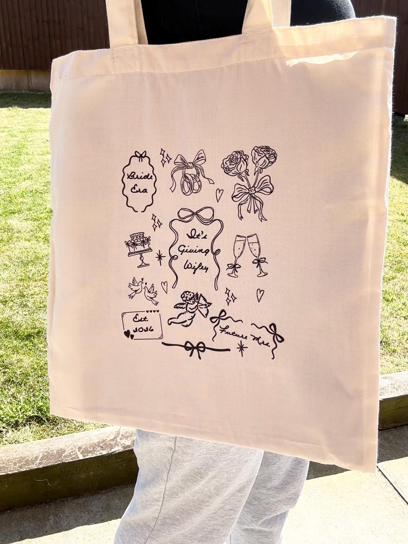 May include: Cream-colored tote bag with black line art illustrations. The design includes text that says "Bride Era", "She's Giving Wifey", and "Est. 2026". Other illustrations include bows, roses, champagne glasses, and a cake.