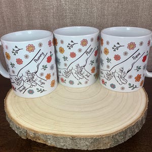 May include: Three white ceramic mugs with floral designs and hand-drawn illustrations of intertwined fingers. The mugs are labeled "Mummy", "Mum", and "Nana".