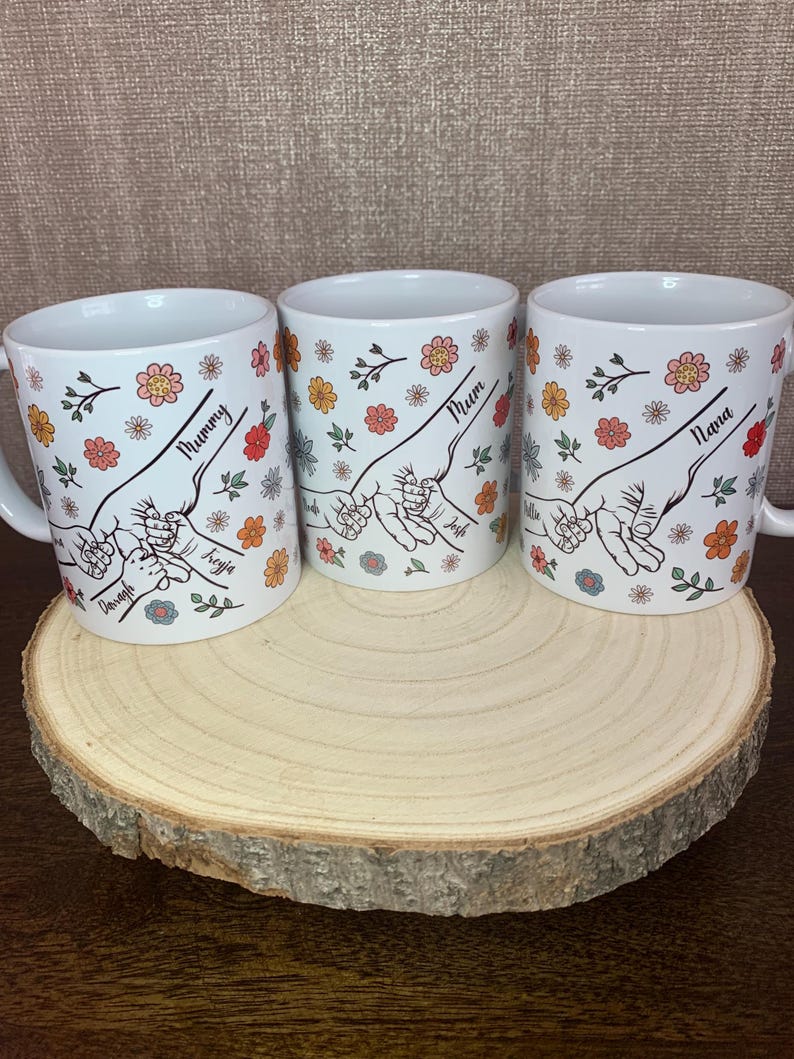 May include: Three white ceramic mugs with floral designs and hand-drawn illustrations of intertwined fingers. The mugs are labeled "Mummy", "Mum", and "Nana".