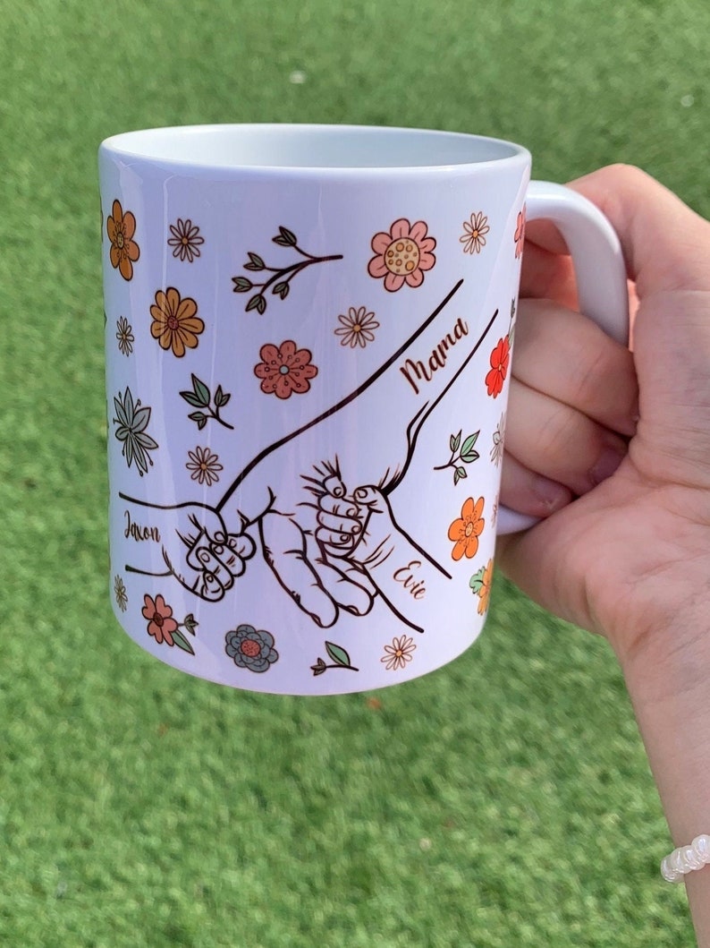May include: White ceramic mug with a floral design and three handprints. The handprints are connected and labeled "Mama", "Jaxon", and "Evie".
