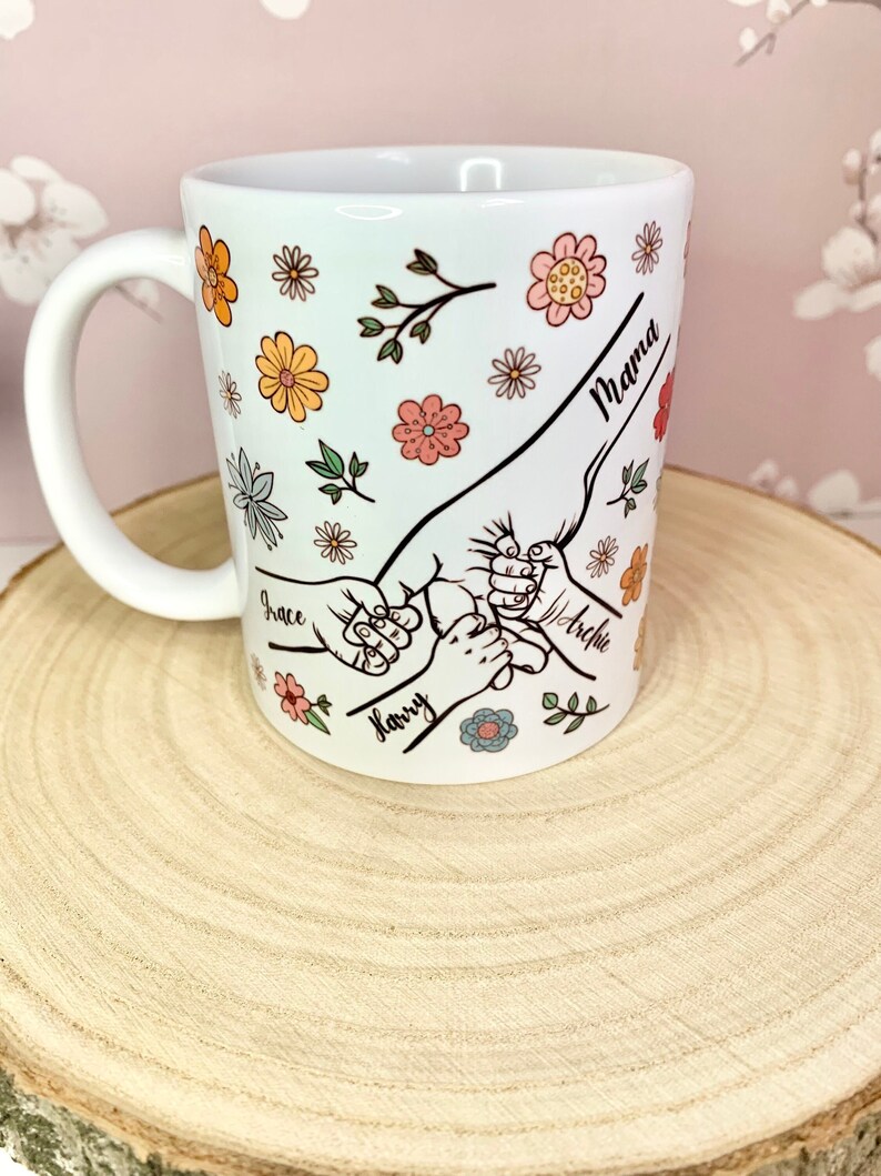 May include: White ceramic mug with a floral design and handprints with the names "Mama", "Grace", "Harry", and "Archie" written on it.