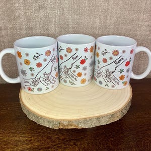 May include: Three white ceramic mugs with floral designs and the words "Mum", "Mum" and "Mama" on them. The mugs are on a wooden stand.