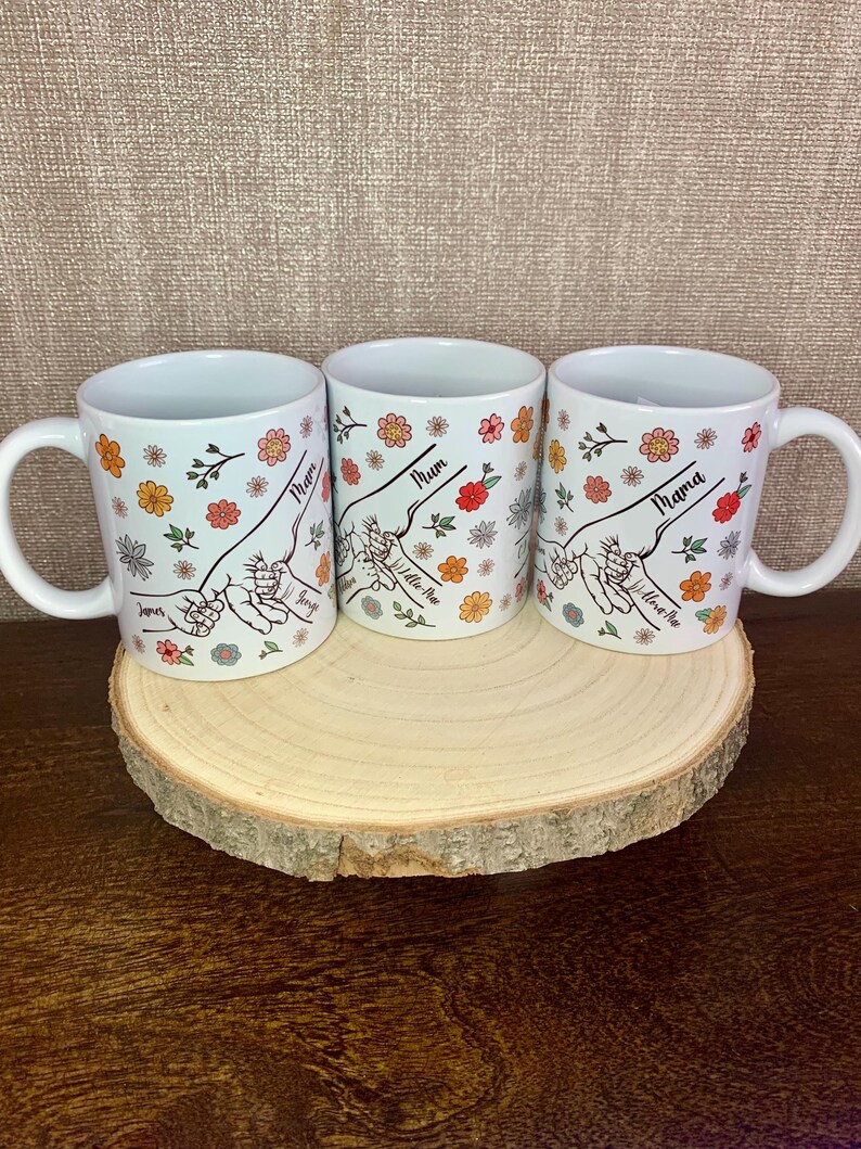 May include: Three white ceramic mugs with floral designs and the words "Mum", "Mum" and "Mama" on them. The mugs are on a wooden stand.