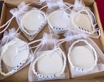 May include: Six white beaded bracelets with names spelled out in letter beads, each in a sheer white gift bag. The names include Izzy, Ruby, Cess, Lex, Tillie, and Liv. The bracelets are displayed in a cardboard box.