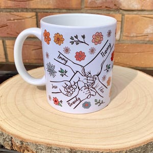 May include: White ceramic mug with a floral design and five hands holding each other. The hands are labeled with the names "Nanny", "Immy", "Jesse", "Beauty", and "Eden".