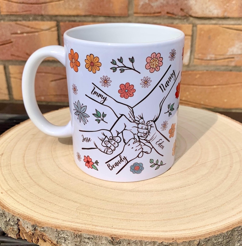 May include: White ceramic mug with a floral design and five hands holding each other. The hands are labeled with the names "Nanny", "Immy", "Jesse", "Beauty", and "Eden".