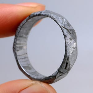Könnte beinhalten: Ein metallischer Ring mit einer dunkelgrauen, texturierten Oberfläche und einem ausgeprägten kristallinen, geometrischen Muster auf dem Band. Der Ring hat einen leicht unregelmäßigen, facettierten Außenrand und eine glatte, polierte Innenseite.