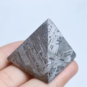 May include: A pyramid-shaped object crafted from metallic meteorite material, displaying a distinct Widmanstätten pattern of intersecting grey and silver crystalline bands across its smooth, geometric faces and sharp, defined edges.