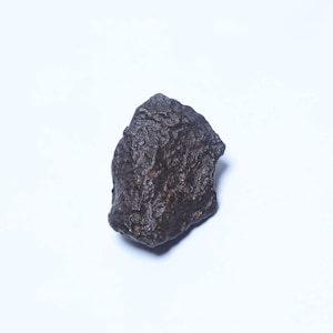 Pode incluir: Uma rocha escura de forma irregular com uma superfície áspera e texturizada. A rocha é de cor castanho escuro e parece ser um espécime de pedra ou mineral natural. O objeto está sobre um fundo branco liso.