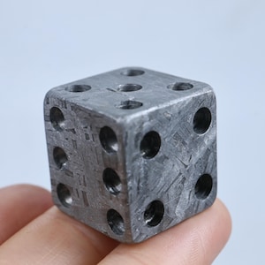 May include: A six-sided die crafted from metallic material with a distinct gray, cross-hatched Widmanstätten pattern across its surfaces. The die features circular, recessed pips arranged in standard numerical configurations on each face.