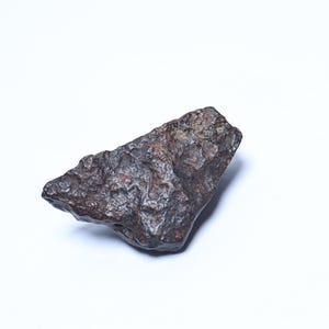 Pode incluir: Uma rocha escura, de forma irregular, com uma superfície áspera e texturizada. A rocha tem uma cor castanha escura e cinzenta, com áreas de oxidação cor de ferrugem. O objeto está sobre um fundo branco liso.