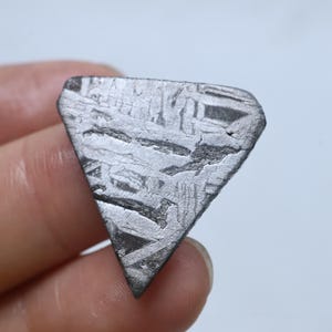 Könnte beinhalten: Ein dreieckiges Metallfragment mit einem ausgeprägten Widmanstätten-Muster aus sich kreuzenden Linien und geometrischen Texturen auf der silbergrauen Oberfläche. Das Objekt hat ein flaches, poliertes Aussehen mit scharfen, definierten Kanten.