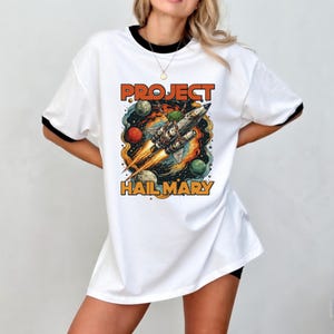 Può includere: T-shirt bianca con bordi neri con una grafica colorata di un razzo spaziale. Le parole "PROJECT" e "HAIL MARY" sono scritte in arancione. Il razzo è circondato da pianeti rossi, verdi e blu.