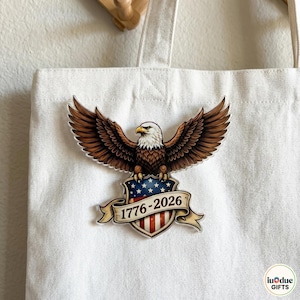 Puede incluir: Un bolso tote blanco con un diseño patriótico. El diseño incluye una ilustración detallada de un águila calva con las alas extendidas sobre un escudo con la bandera estadounidense y el texto "1776-2026".