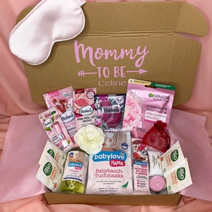 Könnte beinhalten: Eine Geschenkbox für eine werdende Mutter, mit dem rosa Schriftzug "Mommy to Be" und einem Namen. Sie enthält Hautpflegeprodukte, Tee, eine Schlafmaske und eine Kerze. Die Box ist mit rosa Seidenpapier ausgekleidet. Für Celine.