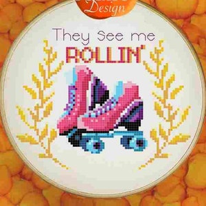 May include: Cross stitch pattern in a white embroidery hoop with a pink and purple roller skate design. The text "They see me rollin'" is above the skate. The hoop is surrounded by orange flower petals on a wood background. The text "Orange Potato Design" is in a bright orange oval above the hoop.