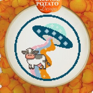 May include: A cross-stitch embroidery hoop with a colorful design of a cow being abducted by a UFO. The design is surrounded by orange flower petals on a white wooden background. The text "ORANGE POTATO Design" is on a bright orange shape above the hoop.