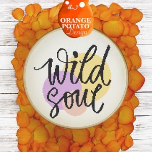 May include: A round embroidery hoop with the phrase "wild soul" in black cursive lettering. The lettering is surrounded by three overlapping circles in shades of purple, peach, and yellow. The hoop is surrounded by orange flower petals on a white wood background.