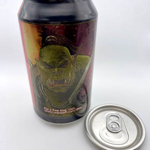 Może przedstawiać: Wysoka, cylindryczna puszka po napoju z czarnym wieczkiem i srebrną pokrywką z zawleczką. Puszka przedstawia grafikę zielonego orka z czerwonymi oczami i mieczem. Tekst na puszce brzmi "For a free trial, visit www.warcraft.com/dew".