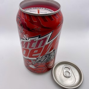 May include: A red and silver Mountain Dew soda can candle with a lit wick. The candle has the Mountain Dew logo and graphics. The can's top is open, and the lid is next to it.