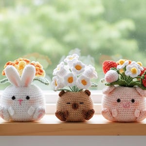 Könnte beinhalten: Drei gehäkelte Tier-Blumentöpfe: ein weißes Kaninchen mit orangefarbenen Blumen, ein brauner Bär mit weißen Blumen und ein rosa Schwein mit Erdbeeren und Blumen. Jeder Topf ist handgefertigt und steht auf einer Holzoberfläche.