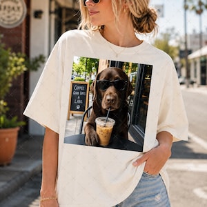 May include: An off-white t-shirt featuring a graphic print of a brown Labrador dog wearing black sunglasses, sitting at a table with an iced coffee. In the background, a chalkboard sign displays the text Coffee Good Days & Labs.