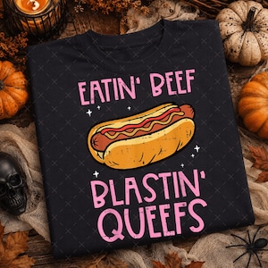 May include: A black crewneck sweatshirt featuring a graphic of a hot dog in the center. The text EATIN' BEEF is printed in pink above the hot dog, and the text BLASTIN' QUEEFS is printed in pink below the hot dog, accented by small white stars.